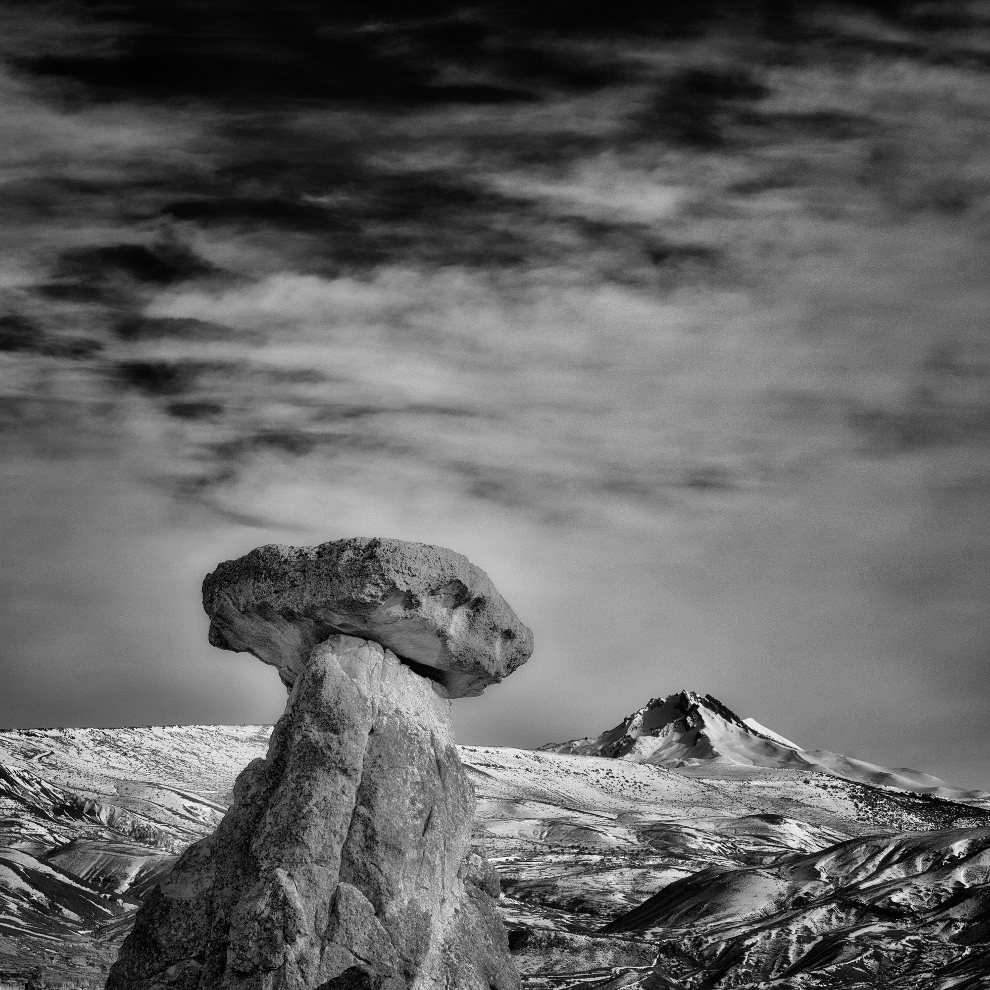 A single broad-capped rock formation in the foreground, with snowy terrain and clouds behind it.